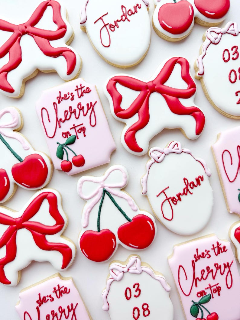 Pu&ograve; includere: Una collezione di biscotti decorati con fiocchi di glassa rossa, targhette rettangolari rosa e bianche con il testo 'she's the Cherry on top', biscotti ovali con il nome 'Jordan' o le date '03 08' in rosso e motivi a ciliegia con fiocchi rosa.