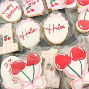 Pu&ograve; includere: Una collezione di biscotti di zucchero confezionati singolarmente con vari disegni, tra cui il testo 'Happy Birthday', 'she's the cherry on top' e 'Halea', insieme a biscotti a forma di numero uno e coppie di ciliegie rosse.