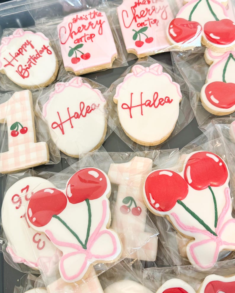 Pu&ograve; includere: Una collezione di biscotti di zucchero confezionati singolarmente con vari disegni, tra cui il testo 'Happy Birthday', 'she's the cherry on top' e 'Halea', insieme a biscotti a forma di numero uno e coppie di ciliegie rosse.