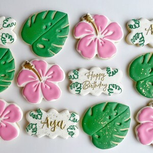 May include: A set of ten decorated sugar cookies featuring a tropical theme. The cookies are decorated with pink hibiscus flowers, green monstera leaves, and gold lettering. The lettering includes "Happy Birthday" and "Aya".