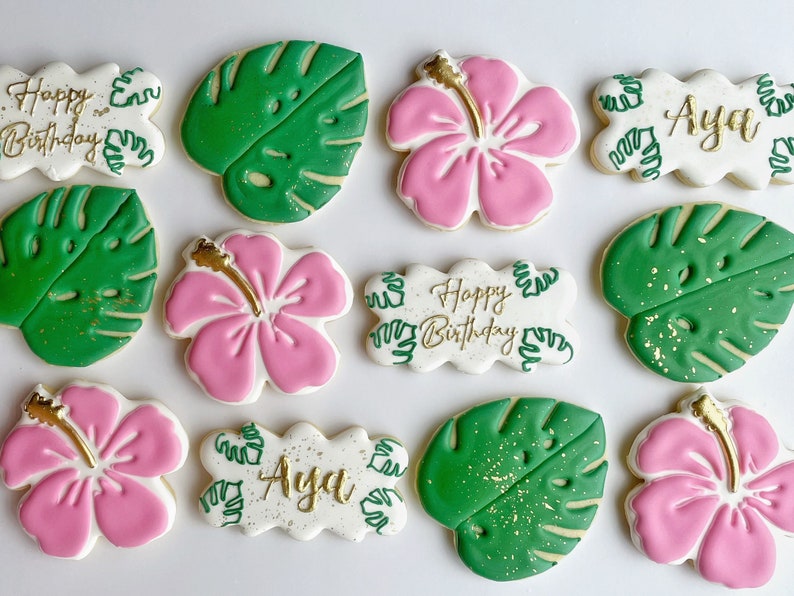 May include: A set of ten decorated sugar cookies featuring a tropical theme. The cookies are decorated with pink hibiscus flowers, green monstera leaves, and gold lettering. The lettering includes "Happy Birthday" and "Aya".