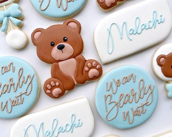 May include: A set of blue and white decorated sugar cookies with the text "We can Bearly Wait" and the name "Malachi". The cookies feature brown teddy bear illustrations.