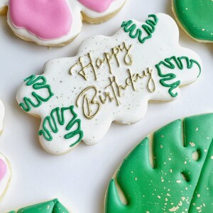 May include: A set of four decorated sugar cookies. One cookie is white with green leaves and the text "Happy Birthday" in gold. Another cookie is pink with a flower design. The remaining two cookies are green with a leaf design and gold sprinkles.