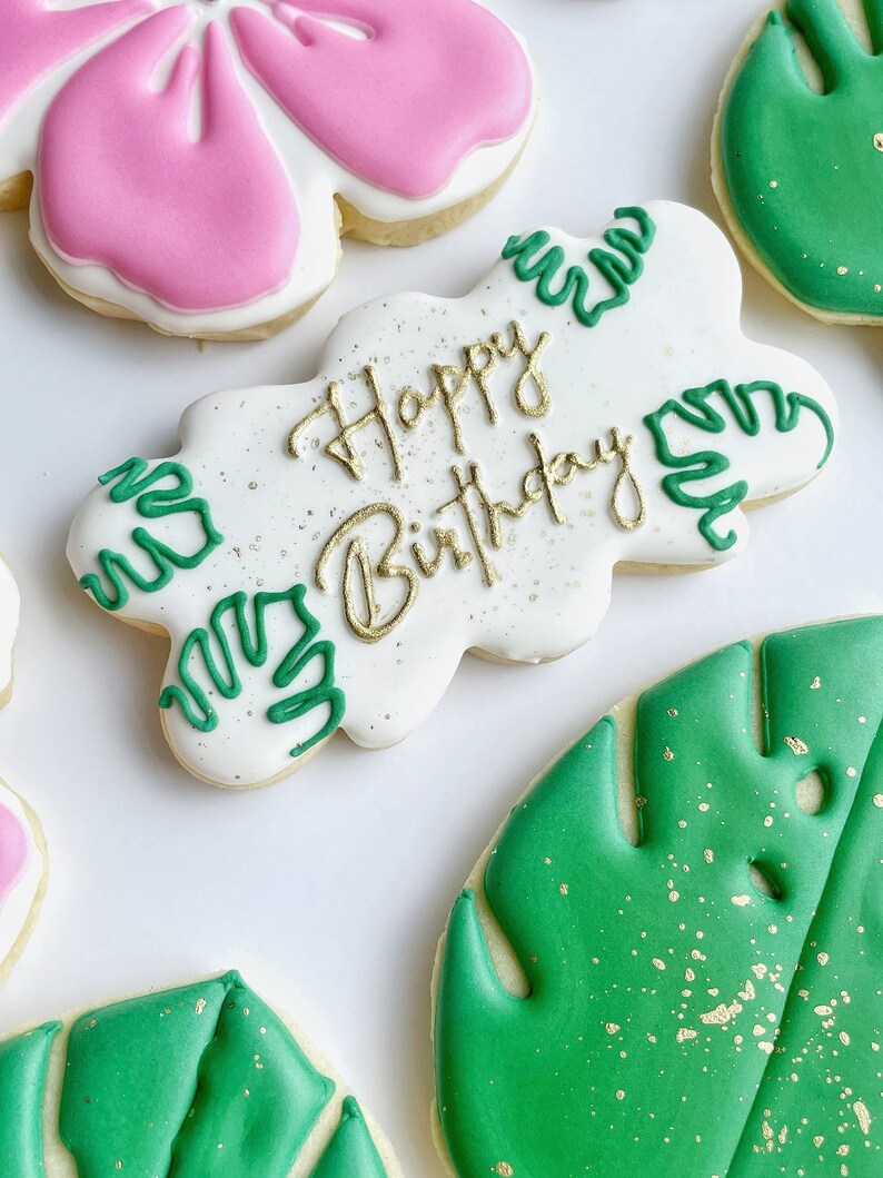 May include: A set of four decorated sugar cookies. One cookie is white with green leaves and the text "Happy Birthday" in gold. Another cookie is pink with a flower design. The remaining two cookies are green with a leaf design and gold sprinkles.
