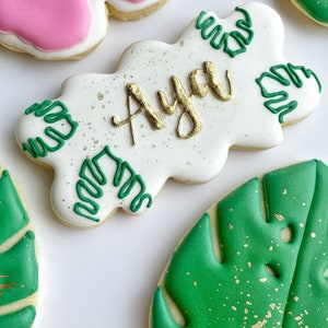 May include: A set of four decorated sugar cookies. One cookie is pink and white with a bow design. Another cookie is white with green and gold leaf designs and the name "Aya" written in gold. The remaining two cookies are green with gold leaf designs.