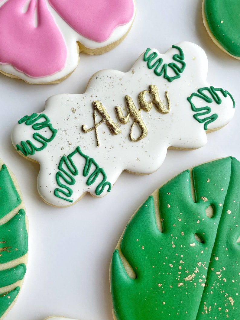 May include: A set of four decorated sugar cookies. One cookie is pink and white with a bow design. Another cookie is white with green and gold leaf designs and the name "Aya" written in gold. The remaining two cookies are green with gold leaf designs.