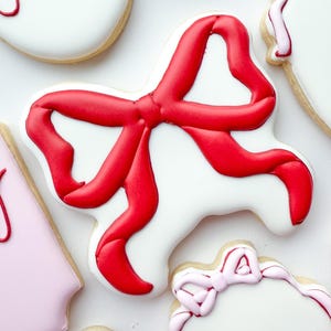 Pu&ograve; includere: Un biscotto a forma di fiocco, decorato con glassa bianca liscia e un design a nastro in glassa rossa vibrante. Il biscotto &egrave; circondato da altri biscotti con glassa bianca e rosa chiaro, alcuni con scritte rosse in corsivo.
