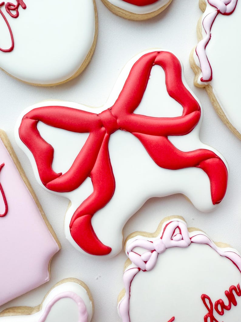 Pu&ograve; includere: Un biscotto a forma di fiocco, decorato con glassa bianca liscia e un design a nastro in glassa rossa vibrante. Il biscotto &egrave; circondato da altri biscotti con glassa bianca e rosa chiaro, alcuni con scritte rosse in corsivo.
