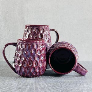 May include: Three ceramic mugs with a textured, bubble-like pattern in shades of purple and burgundy. Each mug has a curved handle and a dark interior. The mugs are arranged on a gray surface.