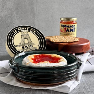 May include: A wheel of Brie cheese with the label "La Bonne Vie" and a jar of "Fromage Ami" Meyer Lemon Wood Orange Pepper Jelly. The cheese is served in a dark teal ceramic dish with handles, topped with red jelly.