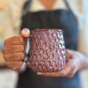May include: A burgundy ceramic mug with a textured, bubbly surface. The mug has a handle and is held in both hands. The mug's exterior has a glossy finish, and the overall aesthetic is rustic and handmade.