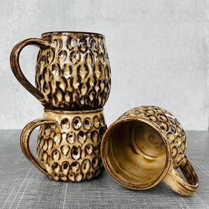 May include: Three brown ceramic mugs with textured, patterned exteriors. The mugs have a dark brown handle and a light brown interior. One mug is lying on its side, revealing the inside. The mugs are stacked and arranged on a gray surface.