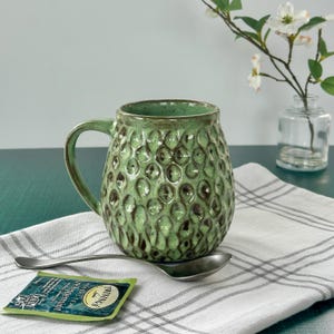May include: A green ceramic mug with a textured, bubble-like pattern. A silver spoon and a tea bag are on a white and gray striped cloth. A small glass vase with white flowers is in the background.