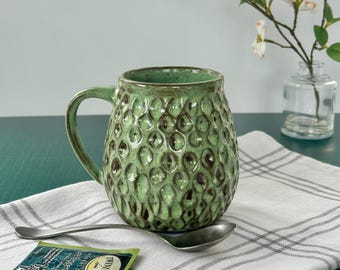 May include: A green ceramic mug with a textured, bubble-like pattern. A silver spoon and a tea bag are on a white and gray striped cloth. A small glass vase with white flowers is in the background.