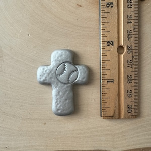 May include: A silver-colored cross-shaped pendant with a baseball design in the center. The pendant is approximately 2.5 inches in length. The cross has a textured surface and is displayed on a wooden background.