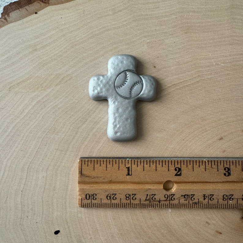May include: A silver-colored cross with a baseball design in the center. The cross has a textured surface and is placed on a wooden background. A wooden ruler is next to the cross.