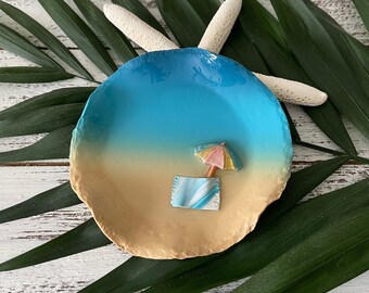 May include: A decorative, round dish with a beach scene. The dish features a blue and tan color scheme, with a miniature beach umbrella and towel. A white starfish rests on top of the dish.