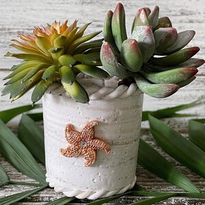 May include: A white cylindrical planter with a textured surface, a rope-like rim, and a copper-colored starfish embellishment featuring a small clear center stone. Two artificial succulent plants are arranged inside, one with green and orange leaves and the other with thick, fleshy green leaves tipped in reddish-pink hues.