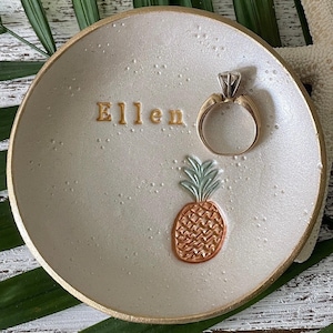 May include: A round, off-white trinket dish with a gold-painted rim, featuring the name Ellen embossed in gold lettering and a small, textured pineapple graphic with a green top and copper-colored base, holding a simple gold metal ring.