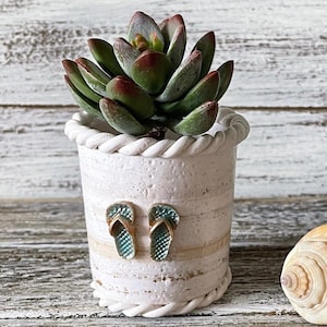 May include: A small, cylindrical white ceramic planter with a textured surface and a rope-like trim at the top and bottom edges. The planter features two small, teal-colored flip-flop embellishments on the front and contains a green succulent.