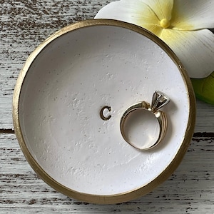 May include: A round white trinket dish with a metallic gold rim, featuring the letter C stamped in the center. A gold ring with a clear stone setting rests inside the dish, which has a textured surface and a slightly uneven, hand-crafted finish.