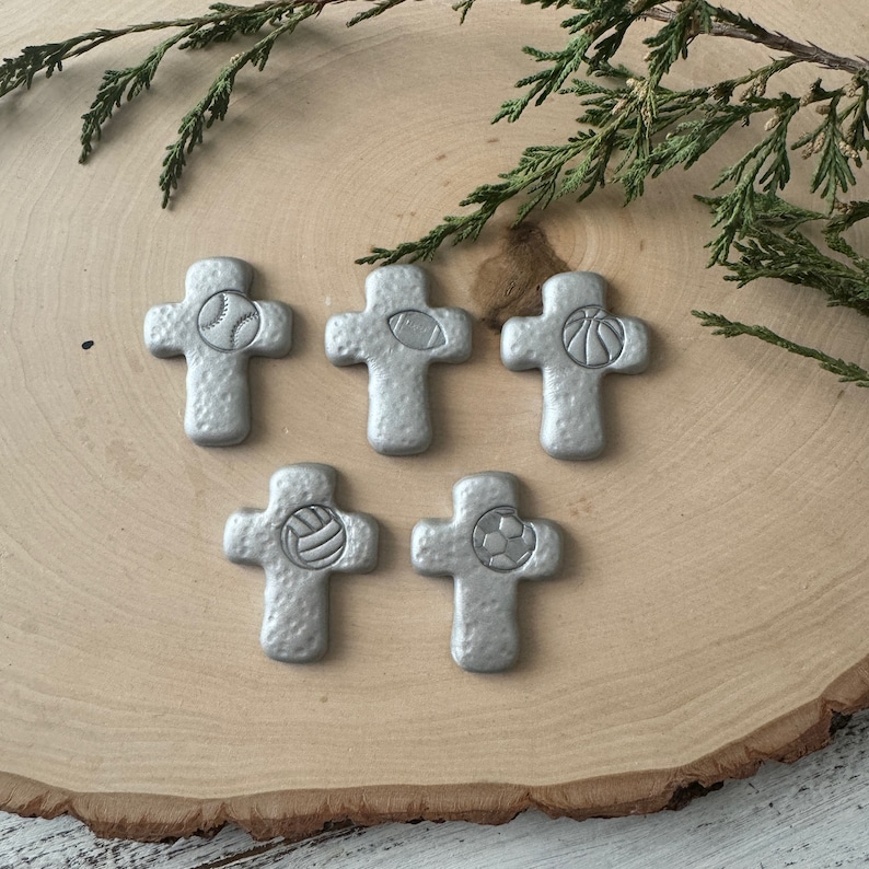 May include: Five silver-colored crosses with sports ball designs: baseball, football, basketball, volleyball, and soccer. The crosses are arranged on a light brown wooden surface, with green sprigs in the background.