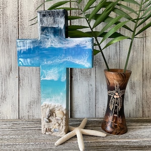 May include: A wooden cross with a blue and white resin ocean wave design. The cross is decorated with seashells and sand. The cross is sitting on a wooden surface with a starfish in front of it.