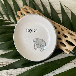 May include: White ceramic trinket dish with a silver manatee design and the name "Taylor" written in silver lettering.