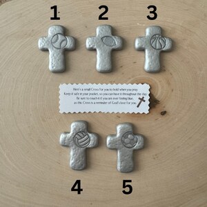 May include: Five silver-colored crosses with sports ball designs: baseball, football, basketball, volleyball, and soccer. A small card reads, "Here's a small Cross for you to hold when you pray..." on a wooden surface.