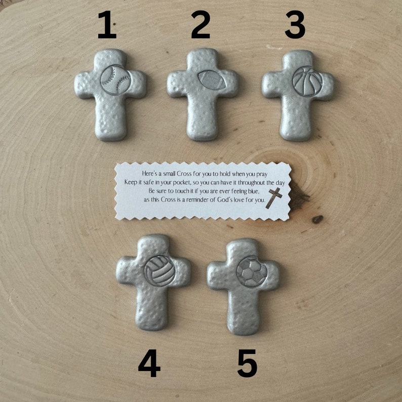 May include: Five silver-colored crosses with sports ball designs: baseball, football, basketball, volleyball, and soccer. A small card reads, "Here's a small Cross for you to hold when you pray..." on a wooden surface.