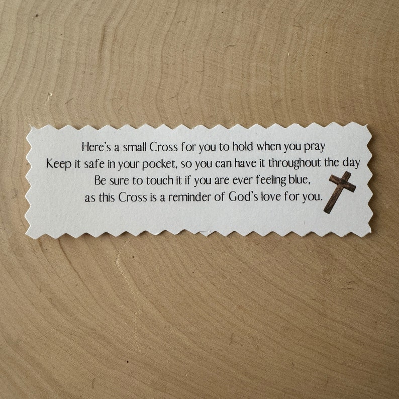 May include: A white paper rectangle with a decorative edge, featuring text and a small cross illustration. The text reads, "Here's a small Cross for you to hold when you pray..." on a light brown wooden surface.