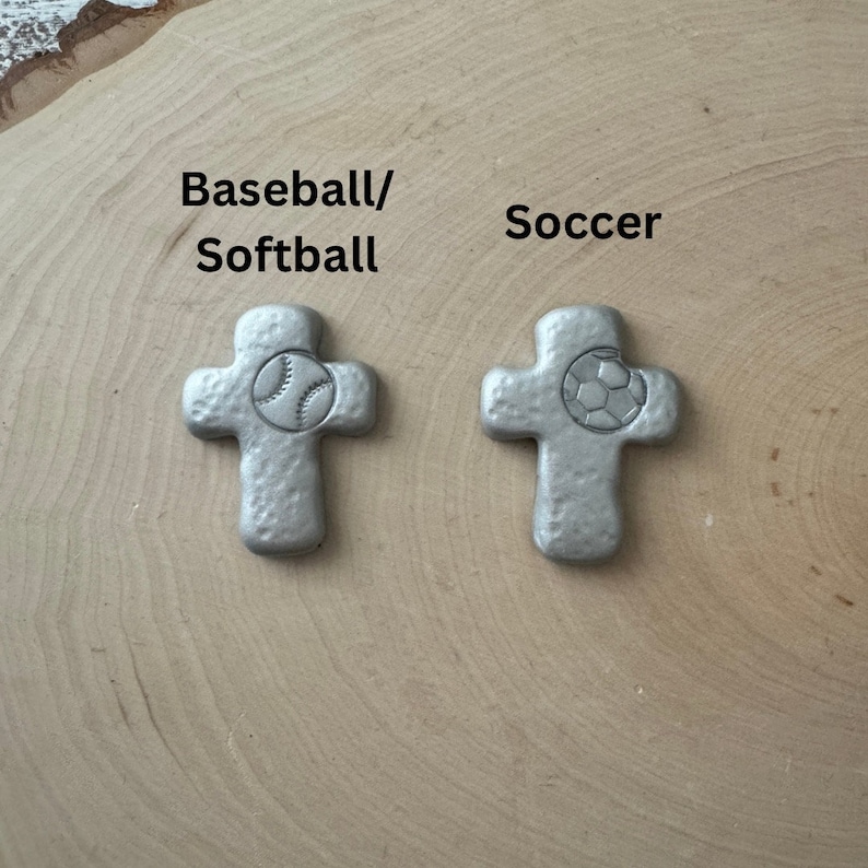 May include: Two silver-colored cross-shaped pendants. One pendant features a baseball, and the other features a soccer ball. The text "Baseball/Softball" and "Soccer" are above the pendants.