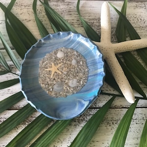 May include: A blue and white resin dish with a swirl design. The dish is filled with sand and seashells, including a starfish.