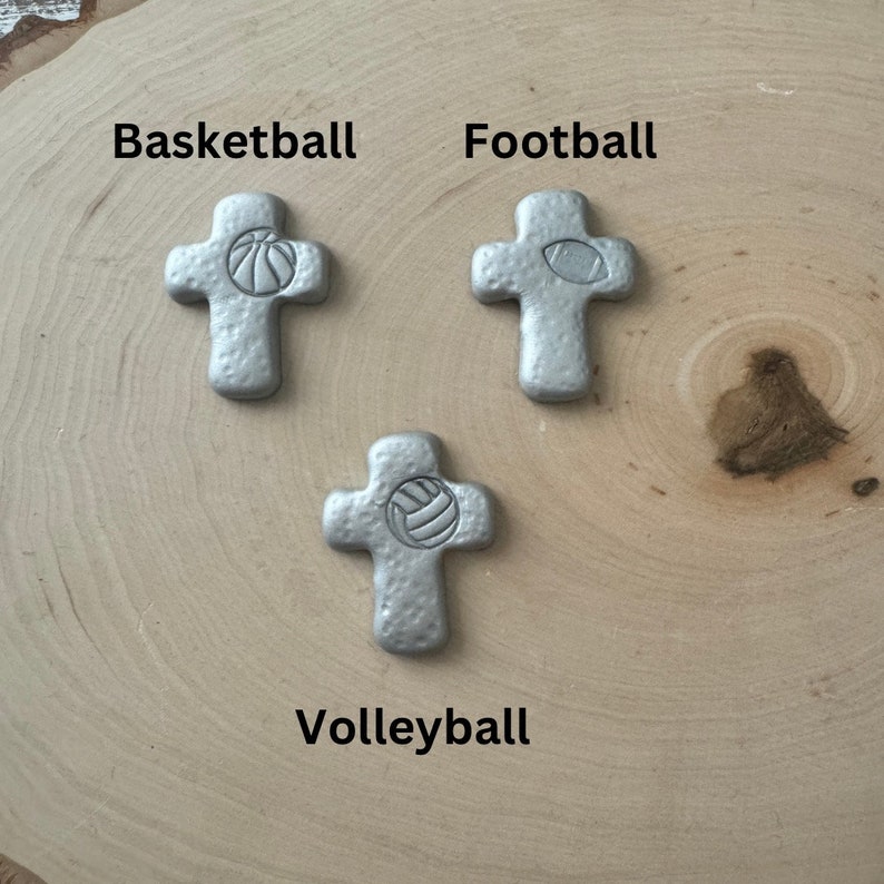 May include: Three silver-colored crosses, each with a different sports ball design: basketball, football, and volleyball. The names of the sports are printed above or below each cross. The crosses are on a light brown, wood-grain surface.