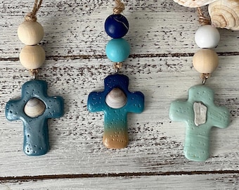 May include: Three hanging cross ornaments made of textured clay. The left cross is dark blue with a small seashell. The center cross features a blue-to-tan gradient with a seashell. The right cross is light green with a small white shell fragment.