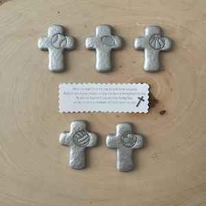 May include: Six silver-colored crosses with sports ball designs: baseball, football, basketball, volleyball, and soccer. A small card with text is included. The crosses are on a light wood surface.