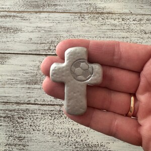 May include: A silver-colored cross with a soccer ball design in the center. The cross has a textured surface and is held in a hand. The background is a light-colored, weathered wood.