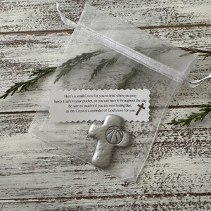 May include: A small, silver-colored cross with a basketball design, presented in a sheer white organza bag. The cross is accompanied by a tag with text about faith and comfort. The background is a rustic, white-washed wooden surface.