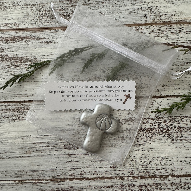 May include: A small, silver-colored cross with a basketball design, presented in a sheer white organza bag. The cross is accompanied by a tag with text about faith and comfort. The background is a rustic, white-washed wooden surface.