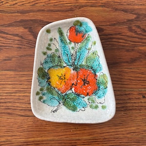 Könnte beinhalten: Kleine, cremefarbene Keramikschale mit floralem Design. Die Blumen sind orange und gelb mit schwarzen Zentren, umgeben von türkisfarbenen und grünen Blättern. Die Schale hat eine unregelmäßige Form.