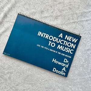 May include: A spiral-bound book titled "A New Introduction to Music" with a dark blue cover. The text includes "Level Two: Pitch & Duration of Tone Using Notation" and the author's name, Dr. Howard A. Doolin. The book is on a light gray surface.