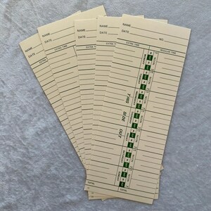 May include: A stack of vintage time cards, each with lines for name, date, and time entries. The cards are a light beige color with green and black printed text. The cards are fanned out, showing the details of the timekeeping layout.