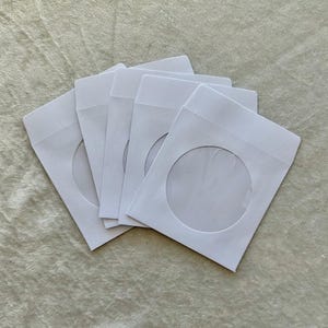 May include: A stack of five white CD/DVD paper sleeves. Each sleeve has a circular window cut out, revealing the disc inside. The sleeves are arranged in a slightly overlapping fan shape.