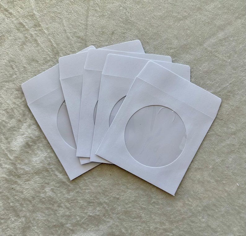 May include: A stack of five white CD/DVD paper sleeves. Each sleeve has a circular window cut out, revealing the disc inside. The sleeves are arranged in a slightly overlapping fan shape.