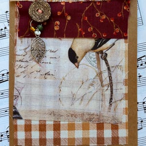 May include: A handmade card with a brown paper background and a layered design of fabric scraps. The top layer features a brown fabric with a floral pattern, followed by a red fabric with a berry pattern, then a white fabric with a bird and handwritten text, and finally a brown and white checkered fabric. A silver leaf charm and a brown button with a red center are attached to the top layer.