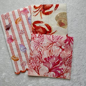 May include: Three paper napkins with a nautical theme. One napkin features red crabs on a white background, another has a red and pink coral design, and the third has red and white stripes with seashells.