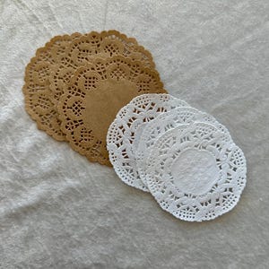 May include: A collection of paper doilies in two colors: tan and white. The doilies have a delicate, lacy design with a circular shape. They are arranged in two separate stacks, showcasing their intricate patterns and textures.