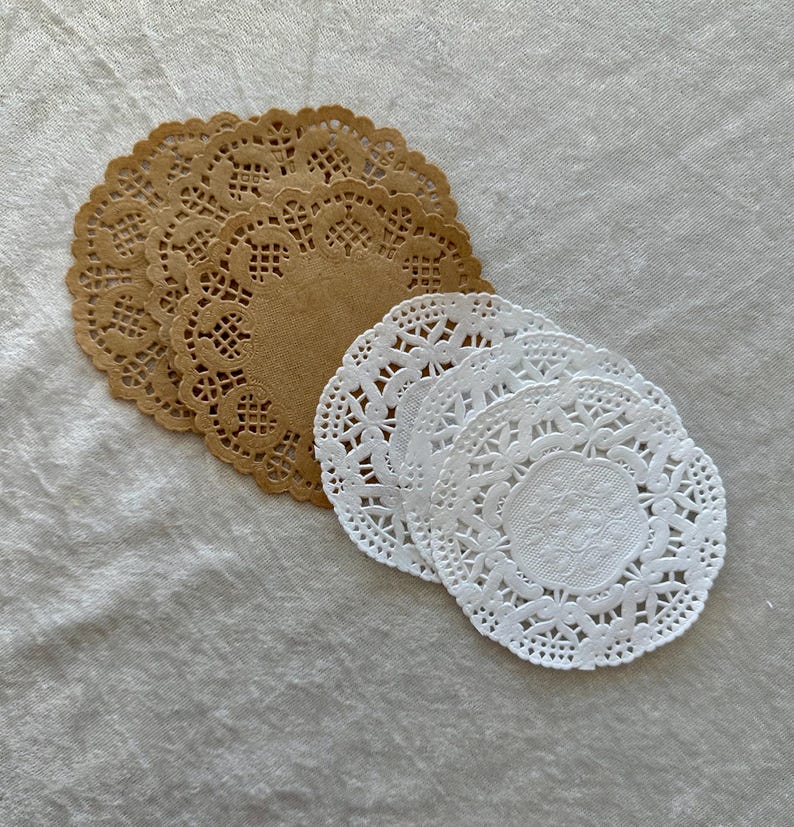 May include: A collection of paper doilies in two colors: tan and white. The doilies have a delicate, lacy design with a circular shape. They are arranged in two separate stacks, showcasing their intricate patterns and textures.
