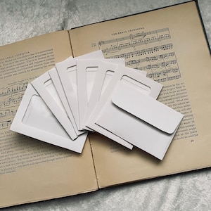 May include: A stack of white envelopes on top of a book with musical notes printed on the pages. The book is open to a page titled "The Small Orchestra".