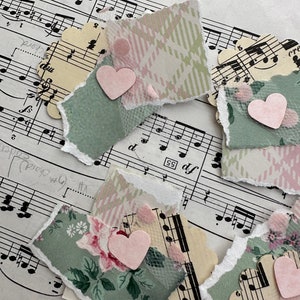May include: A collection of handmade paper embellishments with torn edges, featuring floral, plaid, and musical note patterns. Each piece is adorned with a small pink heart and delicate tulle accents, creating a layered, textured effect. The background is sheet music.